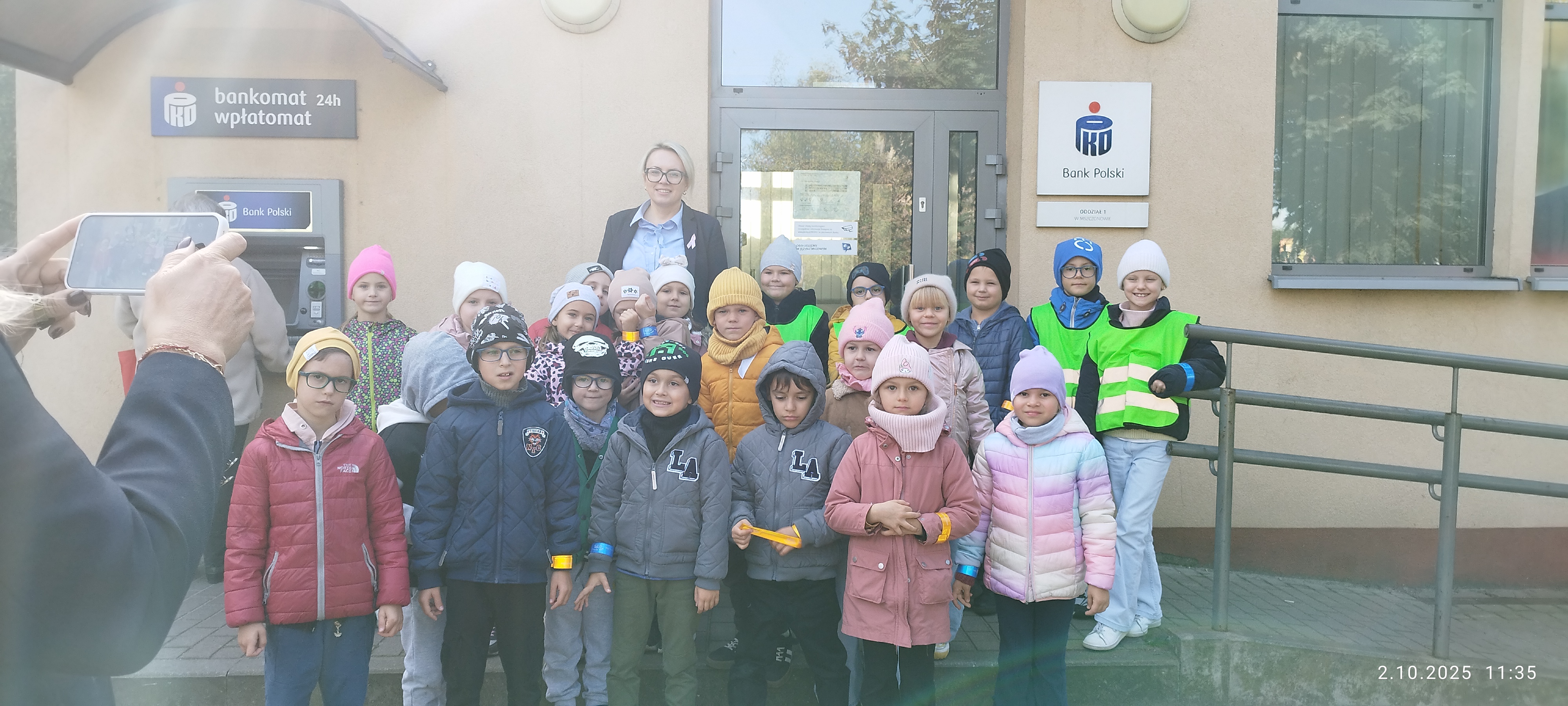 Uśmiechnięci pierwszoklasiści stoją przed bankiem.